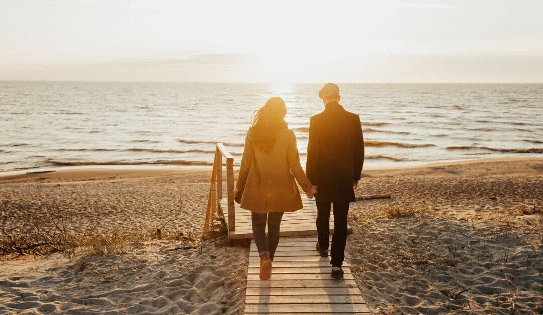 A couple walks down a boardwalk towards sun and ocean