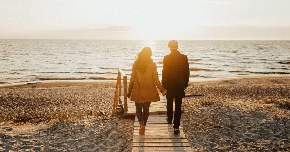 A couple walks down a boardwalk towards sun and ocean