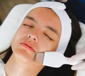 A gloved hand uses a tool on a woman's face to complete a dermasound facial using water and ultrasonic frequency