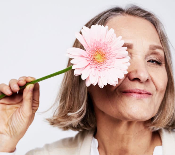 A middle aged woman holds a flower in front of one eye while she smiles at the camera