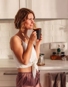 A middle-aged woman stands in her sunlit kitchen and looks off into the distance with a cup of coffee in her hands