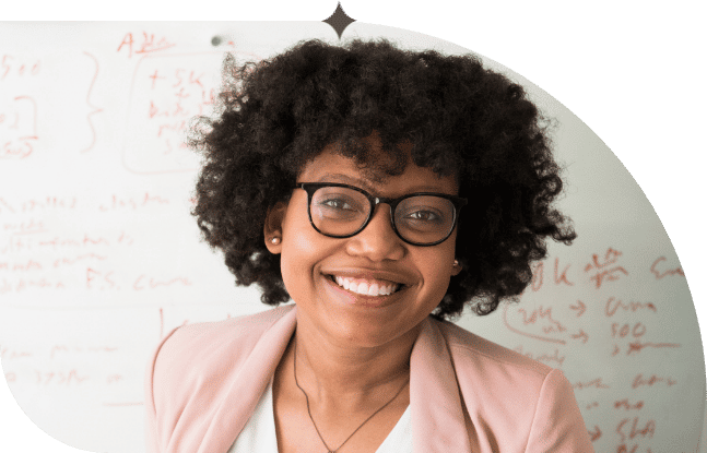 A professional, young African American woman with tight skin in glasses smiles widely at the camera in front of a whiteboard