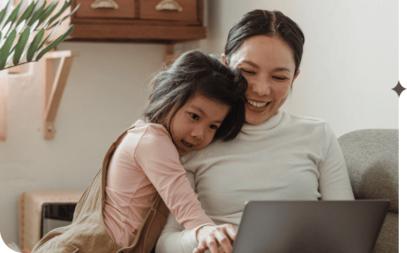 A smiling mother sits on the couch on her laptop, while her daughter embraces her