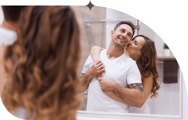 A woman wraps her arms around her husband and kisses him on the cheek as they stand in their bathroom