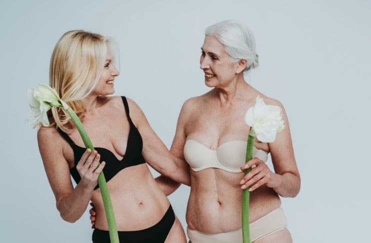 Two older woman in lingerie, each with a flower in their hand, representing their sexual wellness, smile at each other