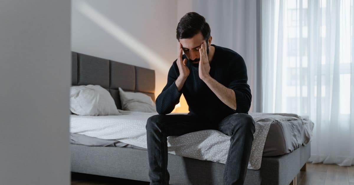 A man sits on the side of his bed fully clothed with his face in his hands