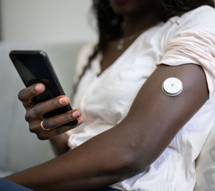 Black woman holding her cellphone and wearing a CGM sensor looks at data from continuous glucose monitoring